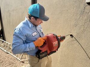 A Holloway Plumbing technician, dressed in uniform, expertly uses a large power tool to drill a hole in an exterior stucco wall. A sturdy ladder and gravel are visible on the ground nearby as part of our professional service setup.